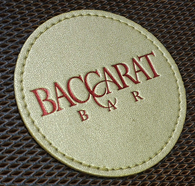 A round, gold vegan leather coaster with the Baccarat Bar logo debossed in metallic red foil.