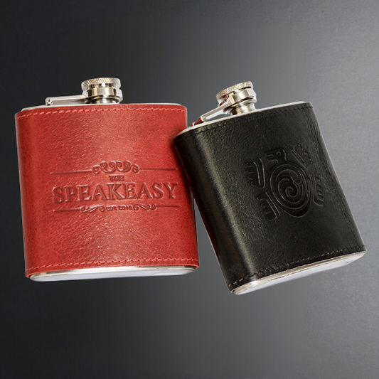 Two leather-look hip flasks on a dark gray gradient background, one in red with 'THE SPEAEASY' embossed on the front and one in black with an embossed spiral logo, both with silver metal flasks visible at the base and caps.