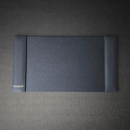 A black blotter with debossed foil decoration, turned edge construction with custom stitching, placed on a textured grey background.