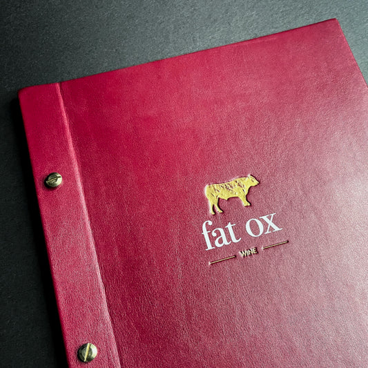 A red faux leather wine list screw post menu with brass posts and a gold debossed logo of a bull's head with the text 'FAT OX WINE' on the cover.