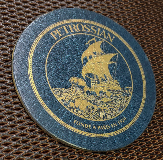 A round, glazed genuine leather coaster with a gold silkscreen deco of a sailing ship on it, set against a brown background.