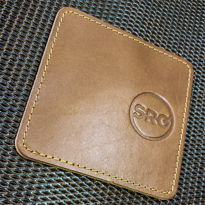 A brown leather coaster with the initials 'SRG' debossed, placed on a textured background.