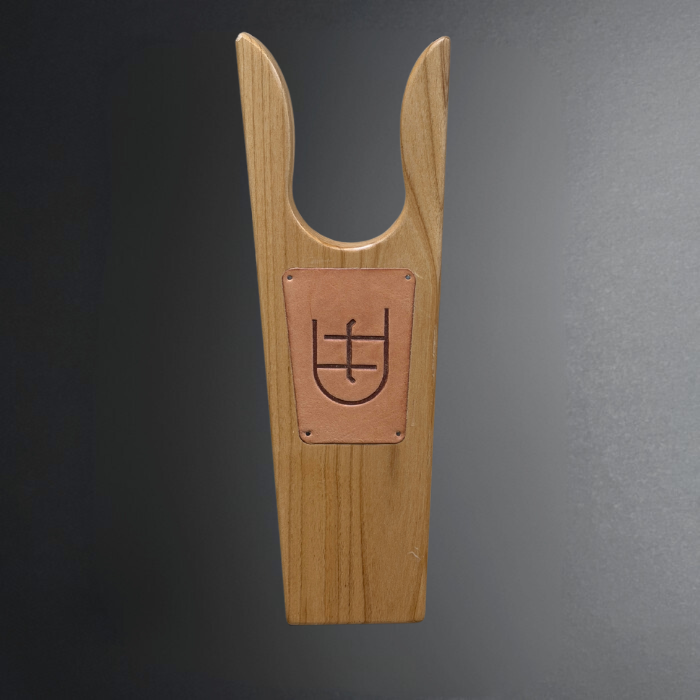 A light natural wood shoe jack with a rectangular copper-colored metal plate in the center featuring a dark engraved symbol, set against a dark gray gradient background.