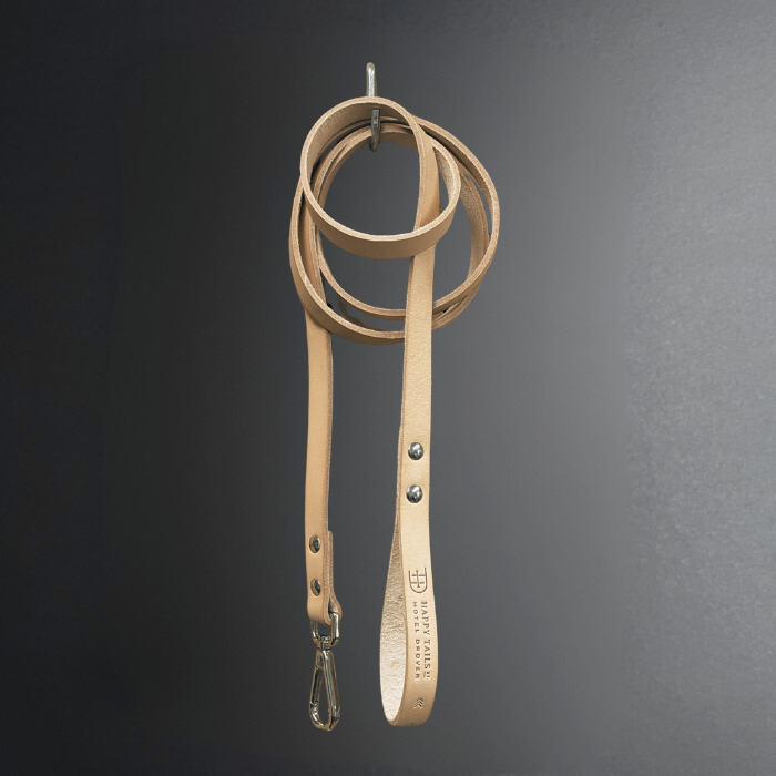 A light tan leather dog leash with silver metal hardware, hanging from a metal ring against a dark gray gradient background. The handle features the brand name 'Happy Tails HOTEL DROVER' and a logo.