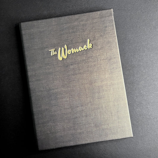 A closed pocket menu cover with 'The Womack' debossed in gold on the front, placed on a dark surface.
